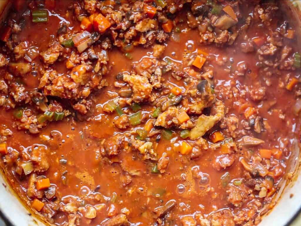 Close-up of a pot of chunky, simmering vegan bolognese with visible pieces of plant-based meat, diced carrots, green herbs, and tomato-based liquid.
